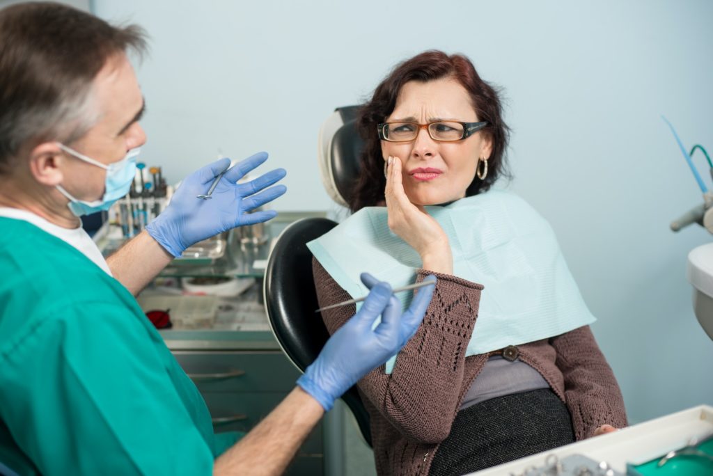 Woman feeling toothache, touching cheek with hand at dental clinic. Senior dentist trying to help