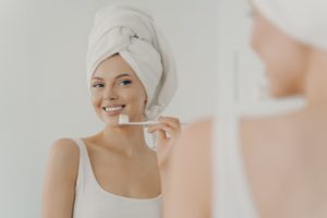 Beautiful young woman with healthy perfect smile brushing teeth and looking in mirror