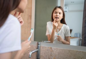 Young Asian woman brushing teeth with sensitive teeth and looking in the mirror.